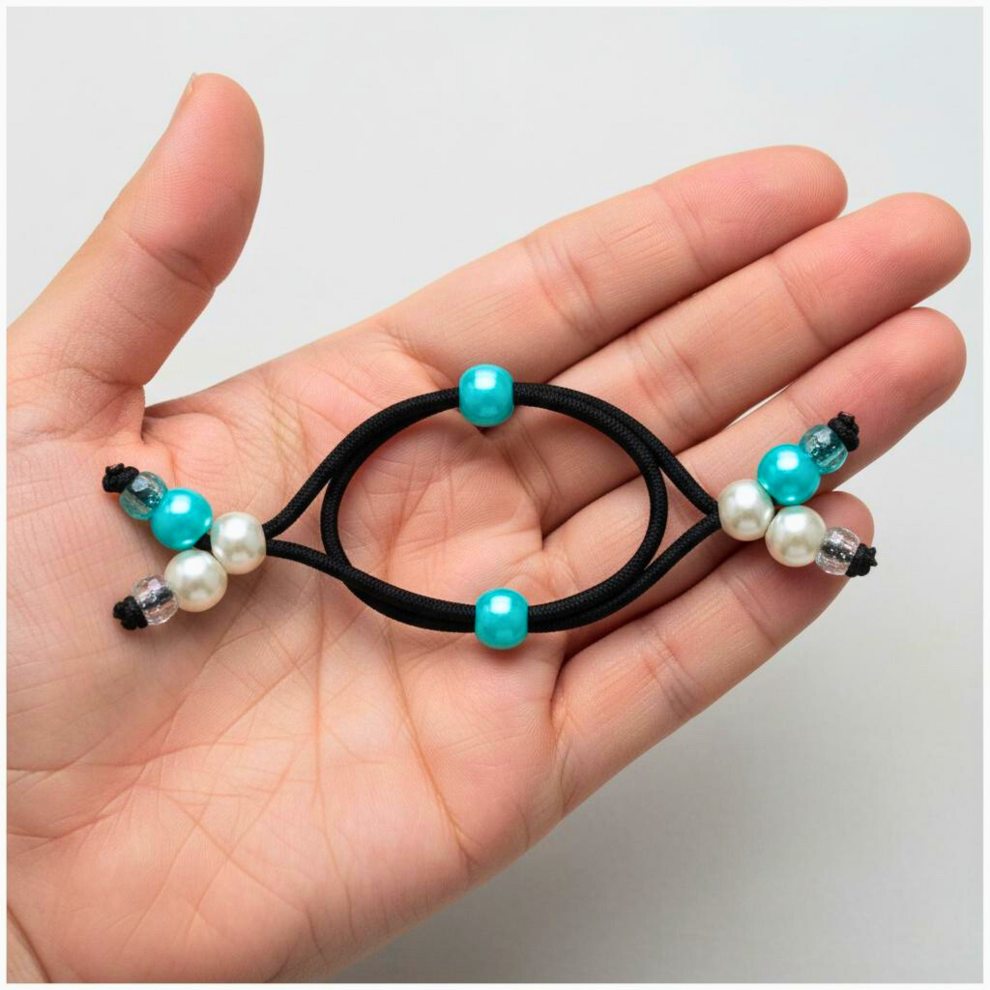 Hand holding a black hair tie with turquoise and pearl beads on a light gray background