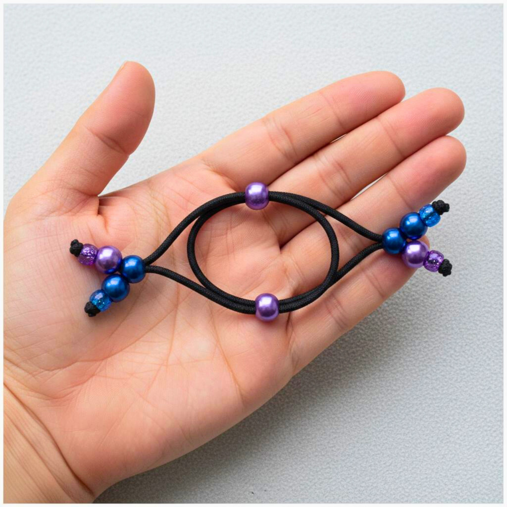 Hand holding black hair tie with purple and blue beads on a light gray background