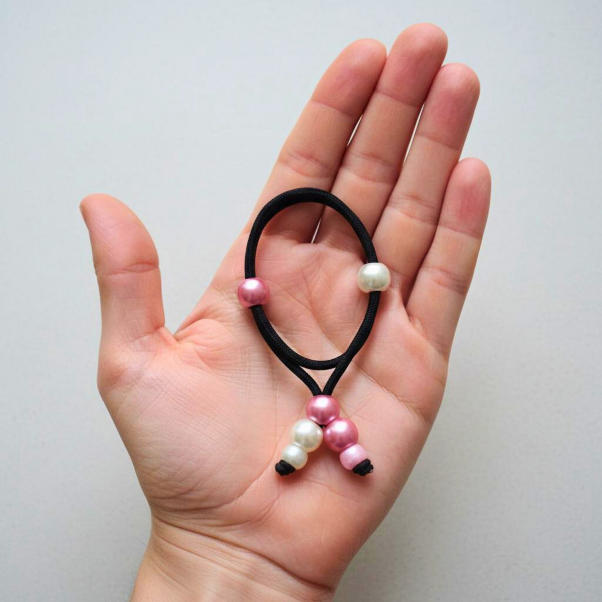 Hand holding a black hair tie with pink and white beads against a light gray background